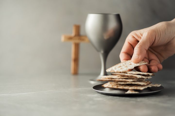 Hands with chalice and communion matzo bread, wooden cross on grey background. Christian communion